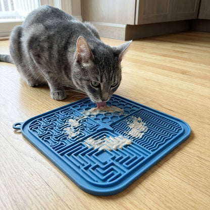 Grey tabby cat licking wet food spread across a blue silicone lick mat on a kitchen floor